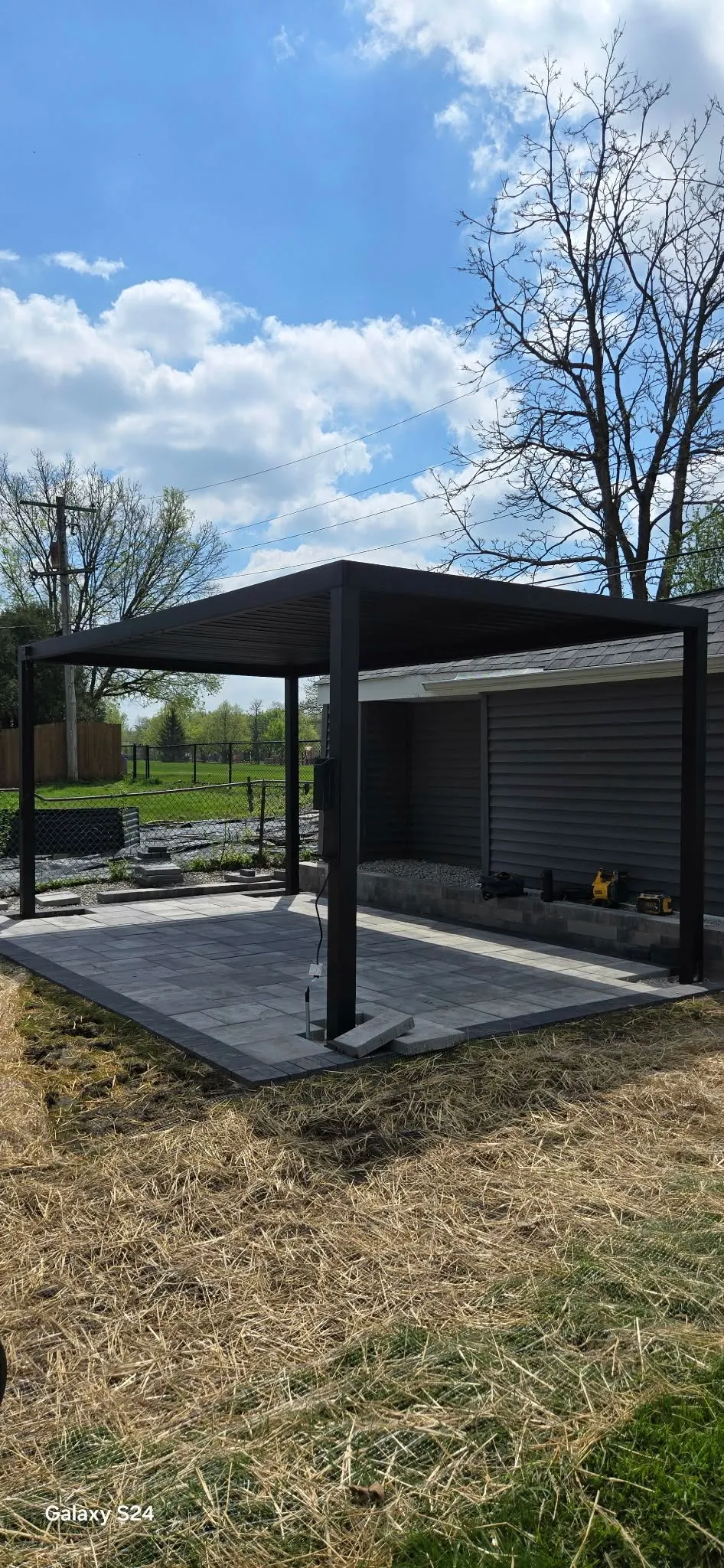 Freestanding black louvered pergola over a newly laid gray paver patio beside a dark-sided garage, with the surrounding yard still under construction