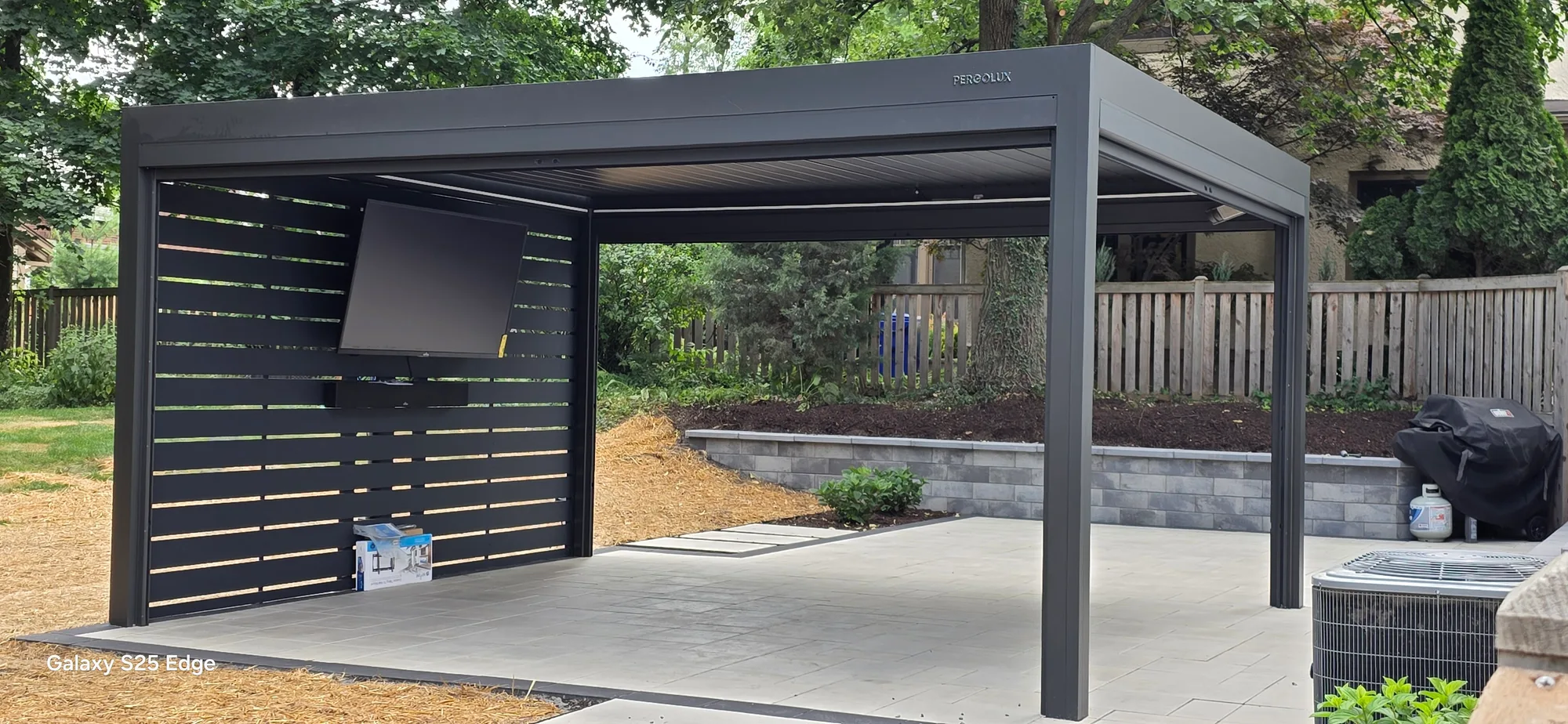 Freestanding black aluminum pergola with a louvered roof and slatted privacy screen, standing over a large-format paver patio in a fenced backyard