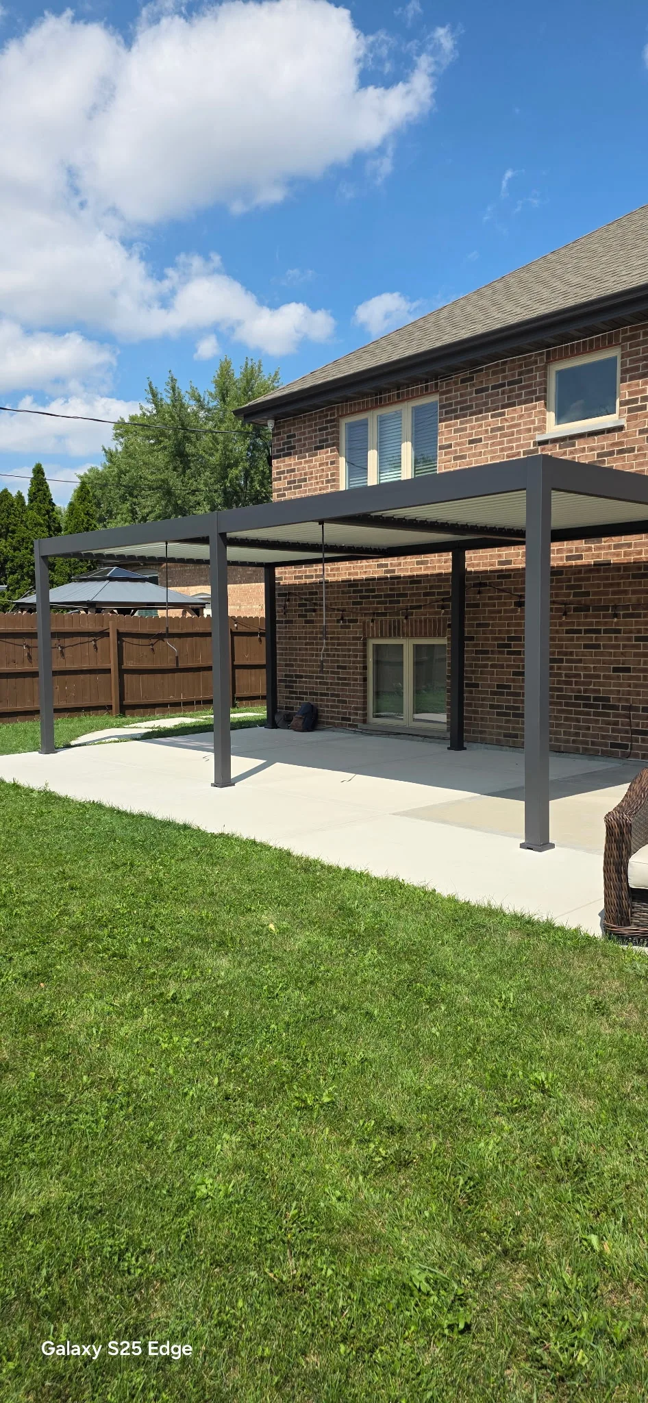 Black aluminum pergola with a louvered roof attached to the rear of a two-story brick home, covering a poured concrete patio