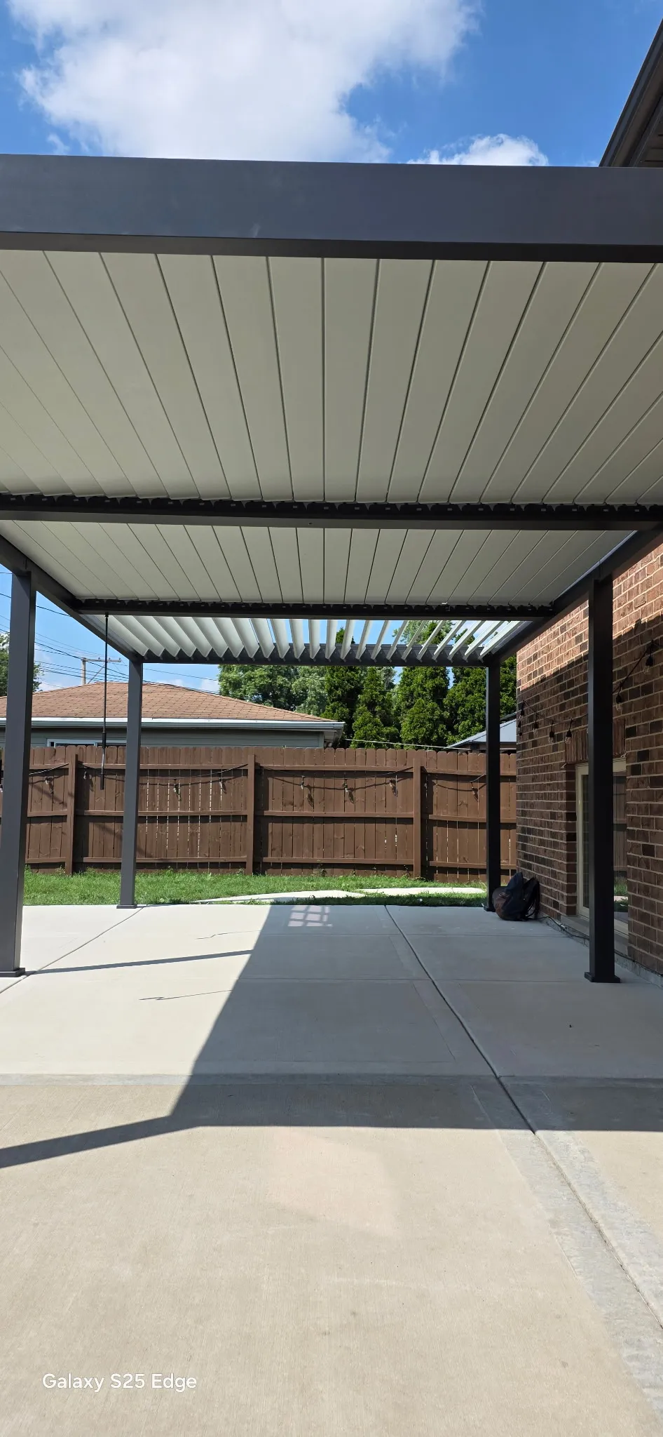 Underside of an attached louvered pergola roof, showing the light-colored louver blades and black steel posts over a poured concrete patio beside a brick home