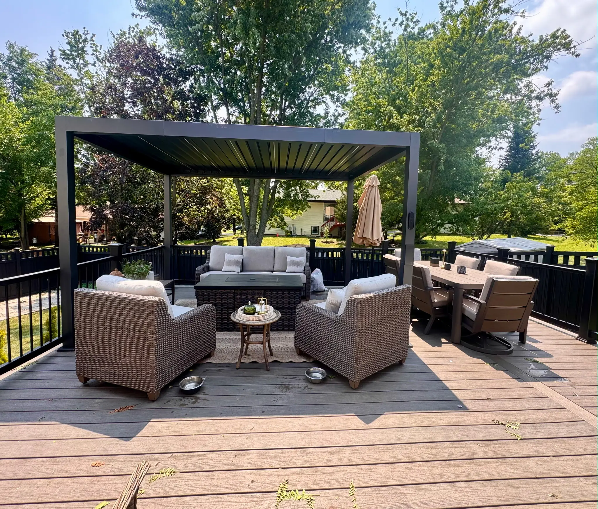 Freestanding black louvered pergola on a composite deck, covering a wicker seating set and an outdoor dining table in a fenced backyard