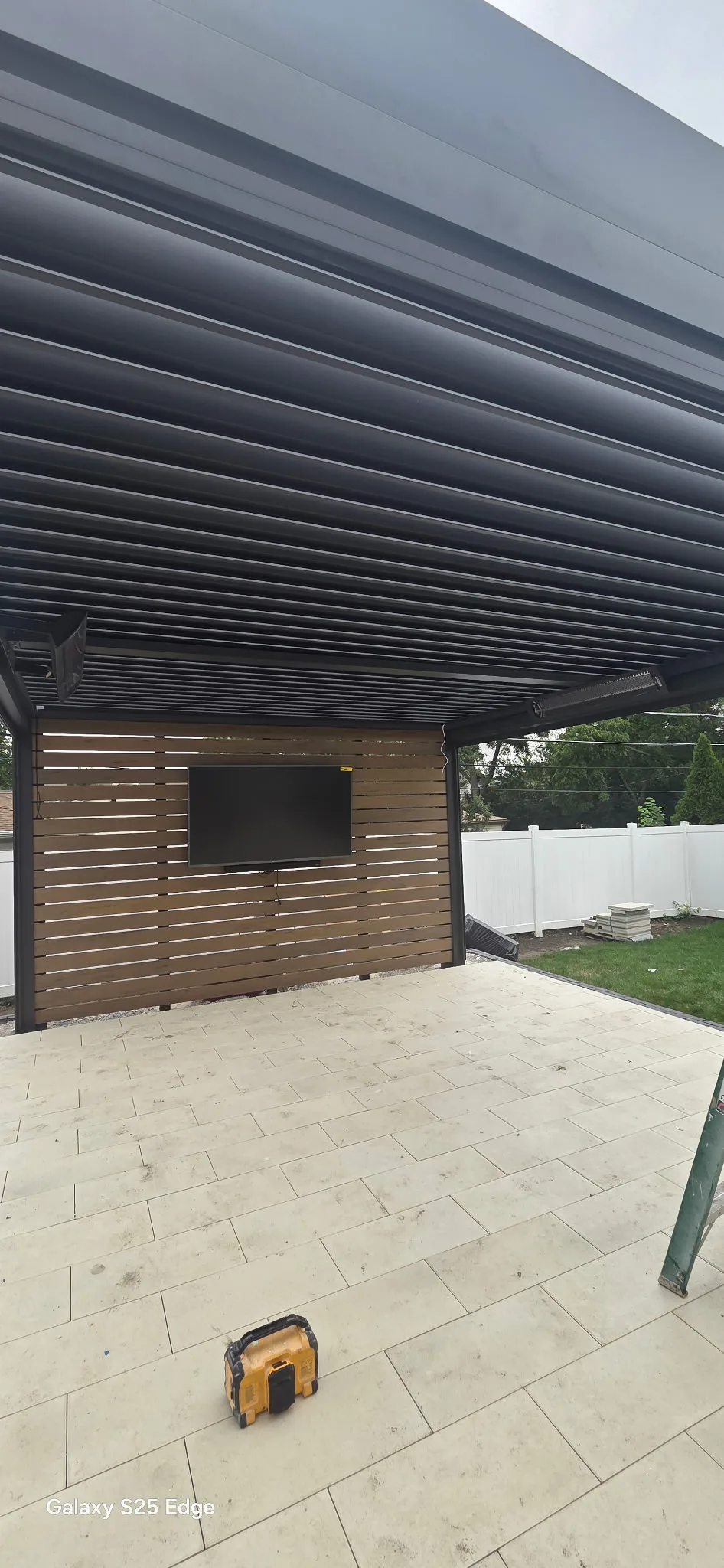 View beneath a closed black louvered pergola roof, with a wood-slat privacy wall, mounted television and large-format paver patio